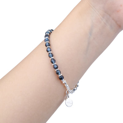 A beaded stretch bracelet with dark blue-gray beads and silver spacer rings, worn on a person's wrist with a dangling silver charm, shown against a plain white background.
